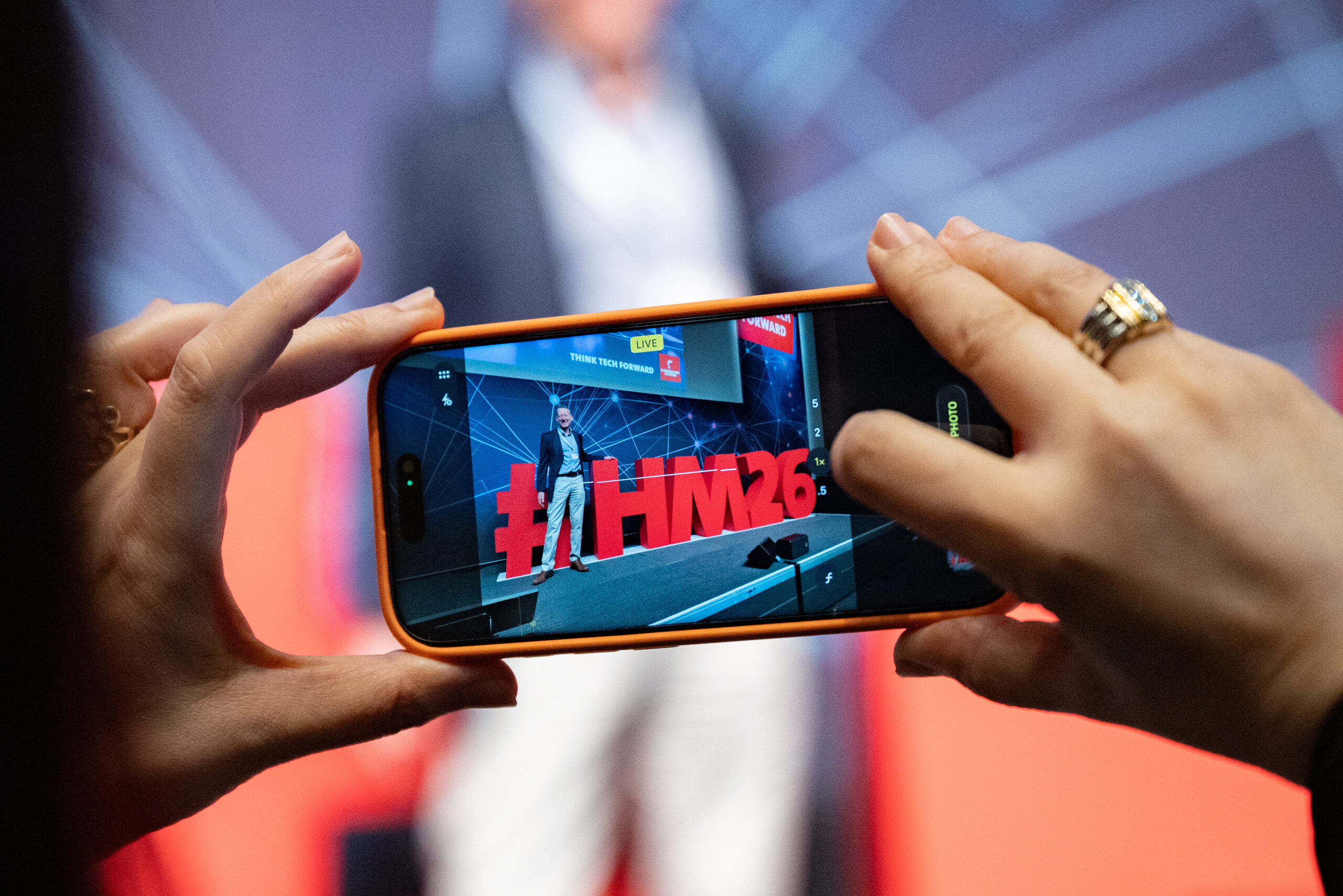 Hannover Messe Preview 2026, Halle 19, Feature mit einem Handy, das auf dem Bildschirm die Bühne mit dem Hashtag HM26 zeigt